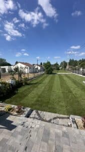 Pelouse rayée impeccable dans un jardin résidentiel ensoleillé. Terrasse pavée, clôture, maisons et ciel bleu nuageux en arrière-plan.