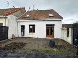 Façade arrière d'une maison blanche en rénovation. Toit tuiles, 2 fenêtres de toit. Patio en dalles et terre. Porte-fenêtre bois et fenêtre. Sac "Appe Chicken".