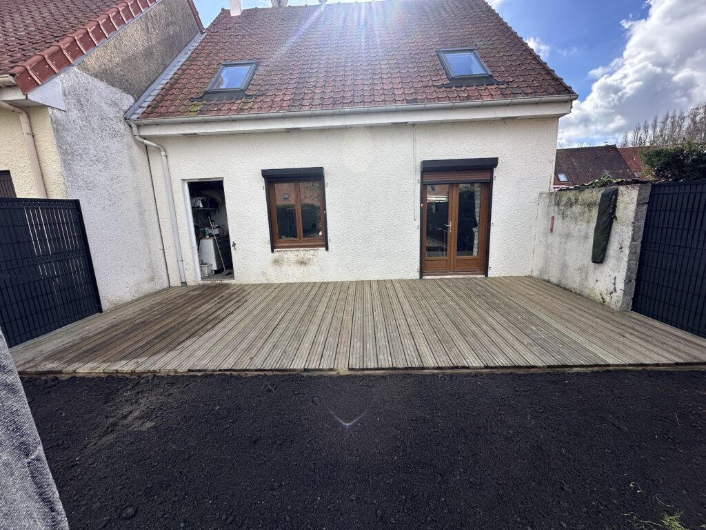Arrière de maison blanche avec toit en tuiles rouges et deux lucarnes. Grande terrasse en bois et jardin en terre noire fraîchement préparée.
